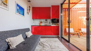 Una sala moderna con un sofá gris, un ventanal grande y una cocina roja al fondo.