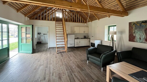 Una habitación amplia con techo alto de madera, una escalera que conduce a un nivel superior, una zona de cocina y un área de estar con una mesa y sillas.