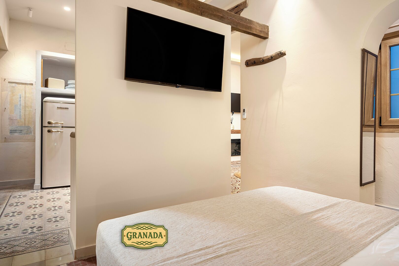 A bedroom with a bed, a television mounted on the wall, a refrigerator, and a wooden beam ceiling.