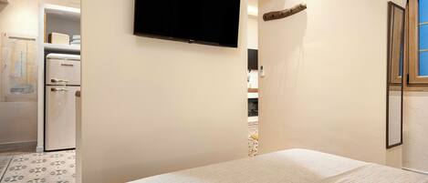 A bedroom with a bed, a television mounted on the wall, a refrigerator, and a wooden beam ceiling.