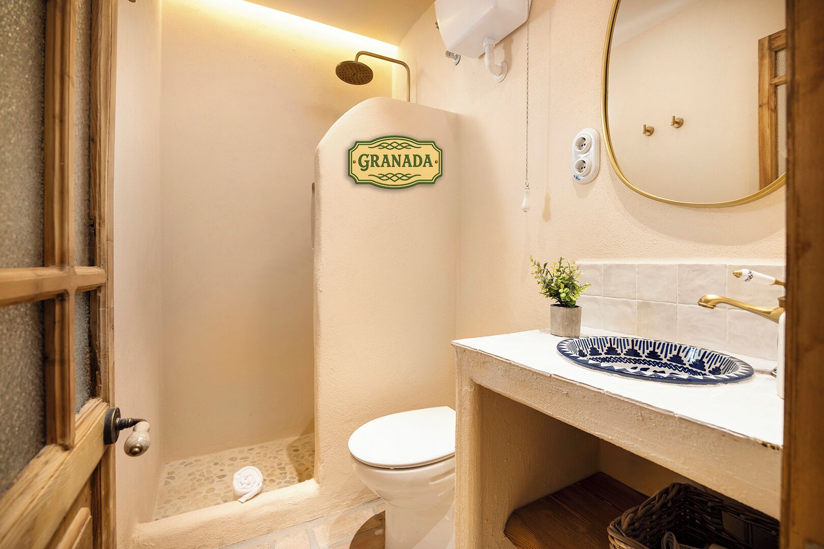 Un baño con inodoro, ducha, lavamanos con un plato decorativo, una planta pequeña en maceta y un espejo montado en la pared.
