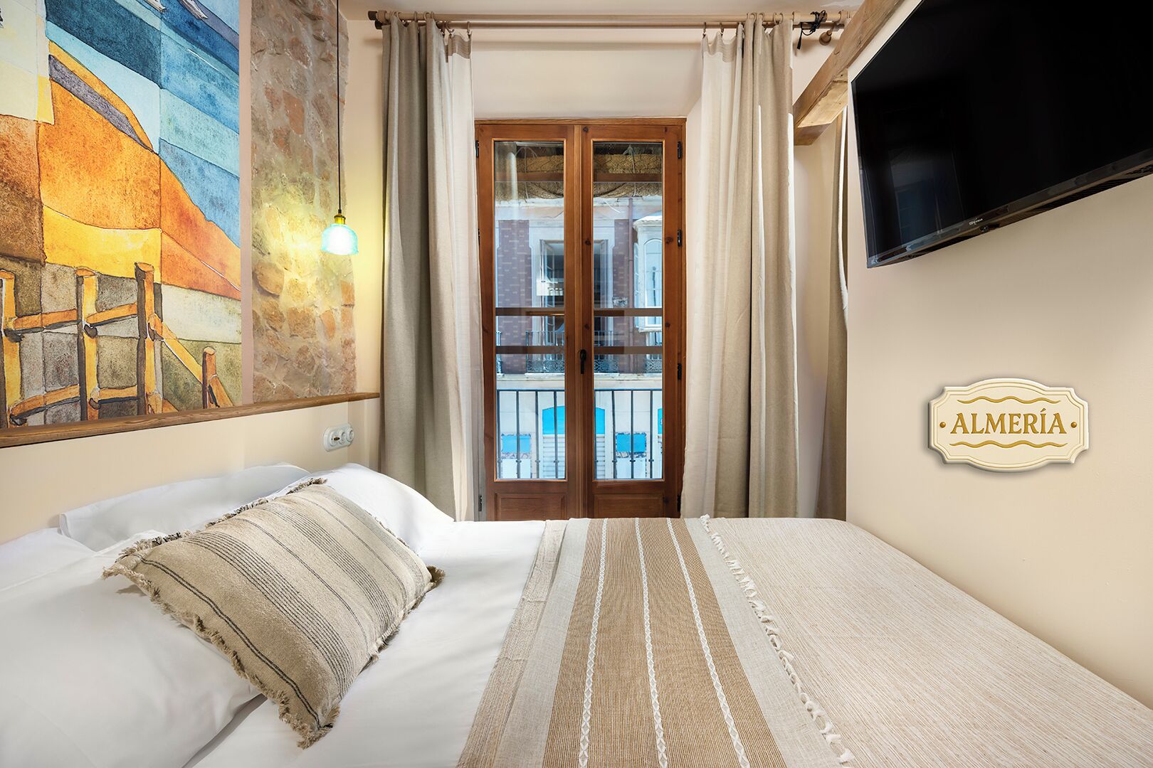 A hotel room with a bed, a painting on the wall, a TV mounted on the wall, and a sign that says "ALMERÍA".