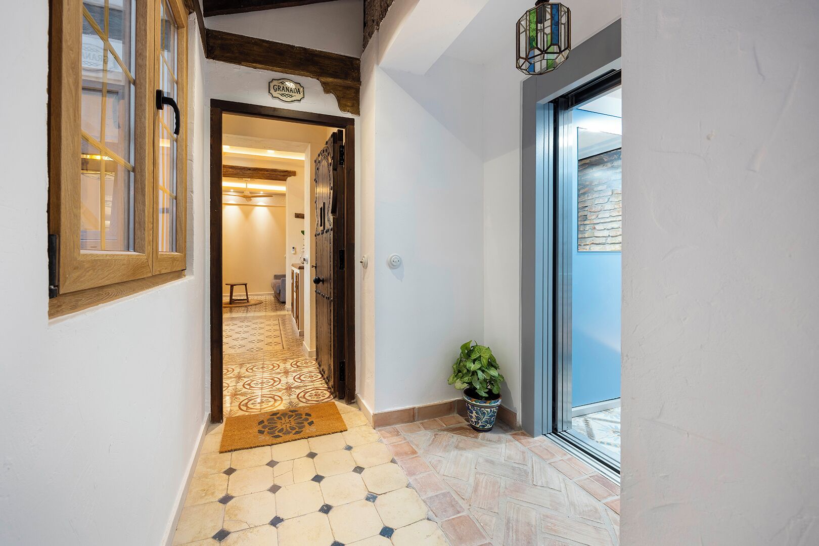 A hallway with patterned tiles, a wooden door, a potted plant, and a view through a glass door.