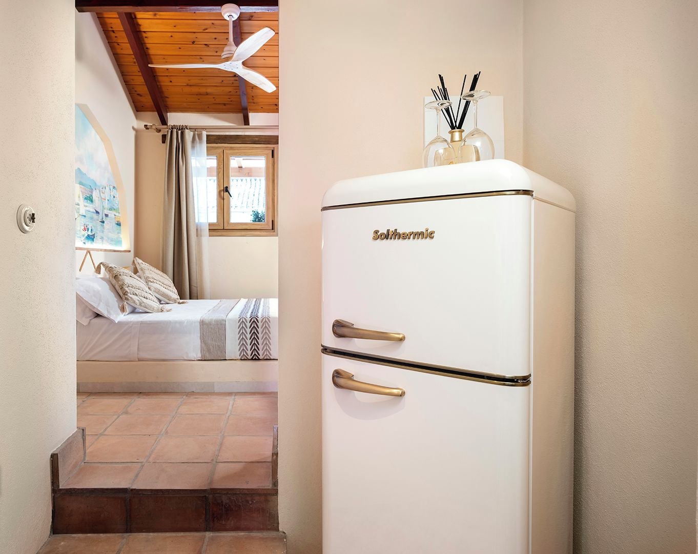 A compact room with a bed, a white refrigerator, and a ceiling fan.