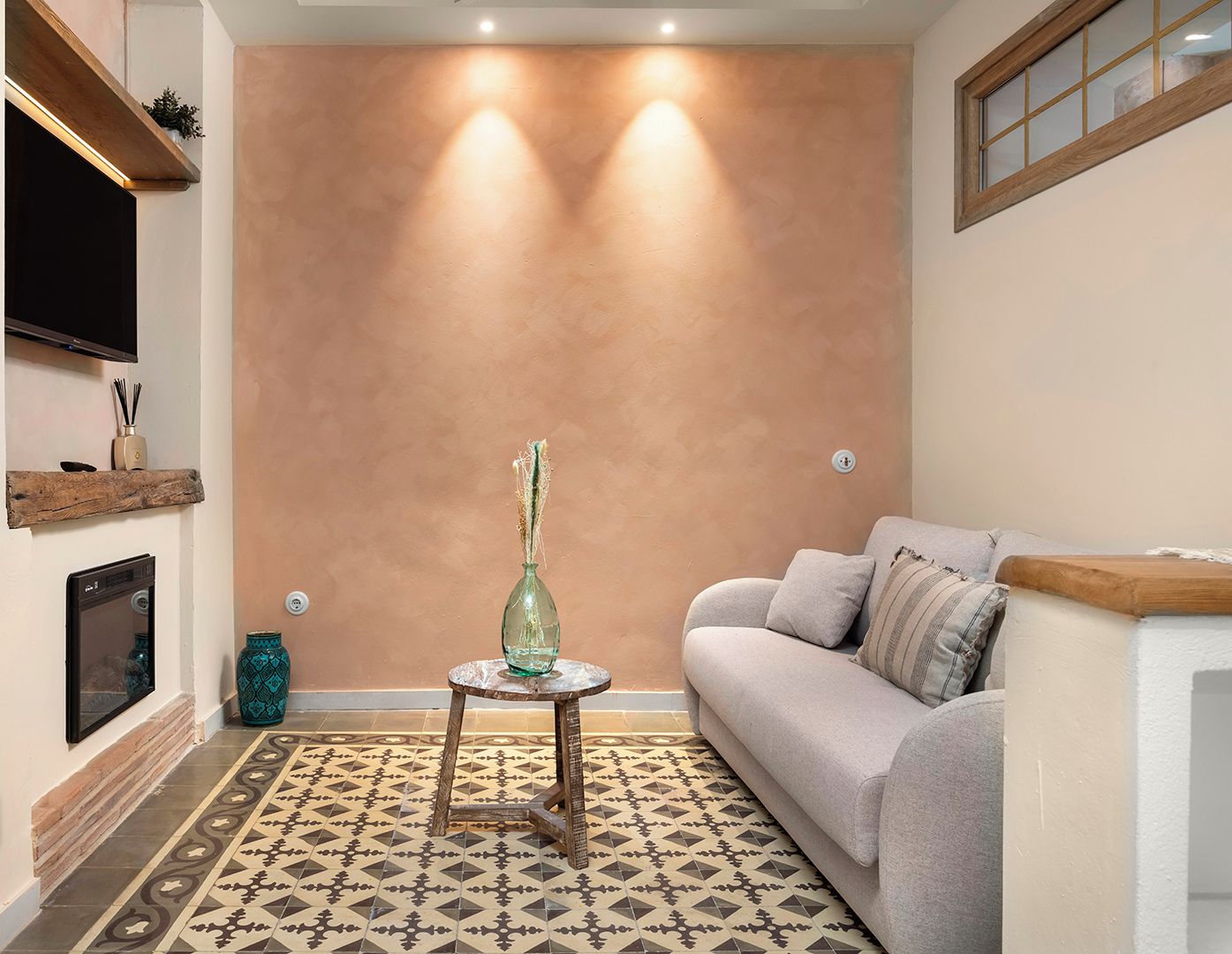 Una sala de estar con un sofá, una mesita redonda con un florero, un televisor montado en la pared y una alfombra estampada.
