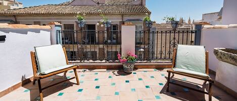 Un balcón con dos sillas de madera, una maceta con flores rosadas y un suelo de baldosas.