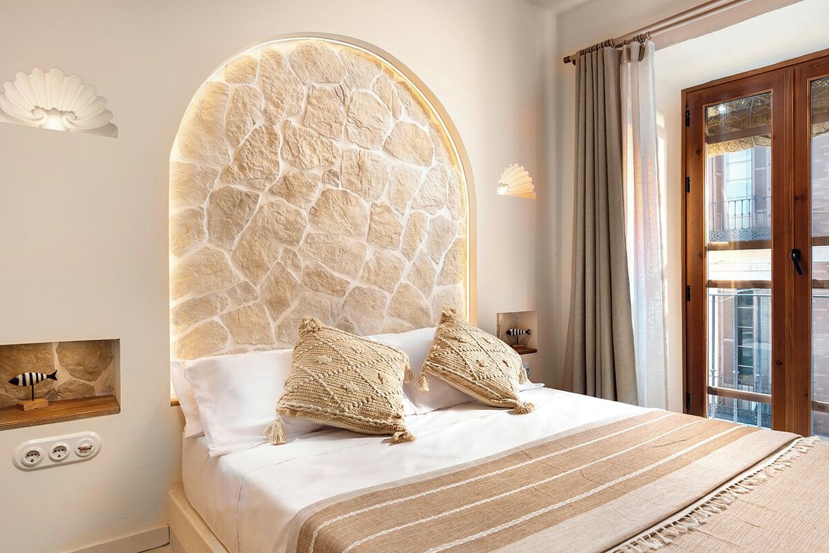 A bedroom with a stone headboard, a bed with striped bedding, a small shelf with a fish decoration, and a view of the outside through a glass door.