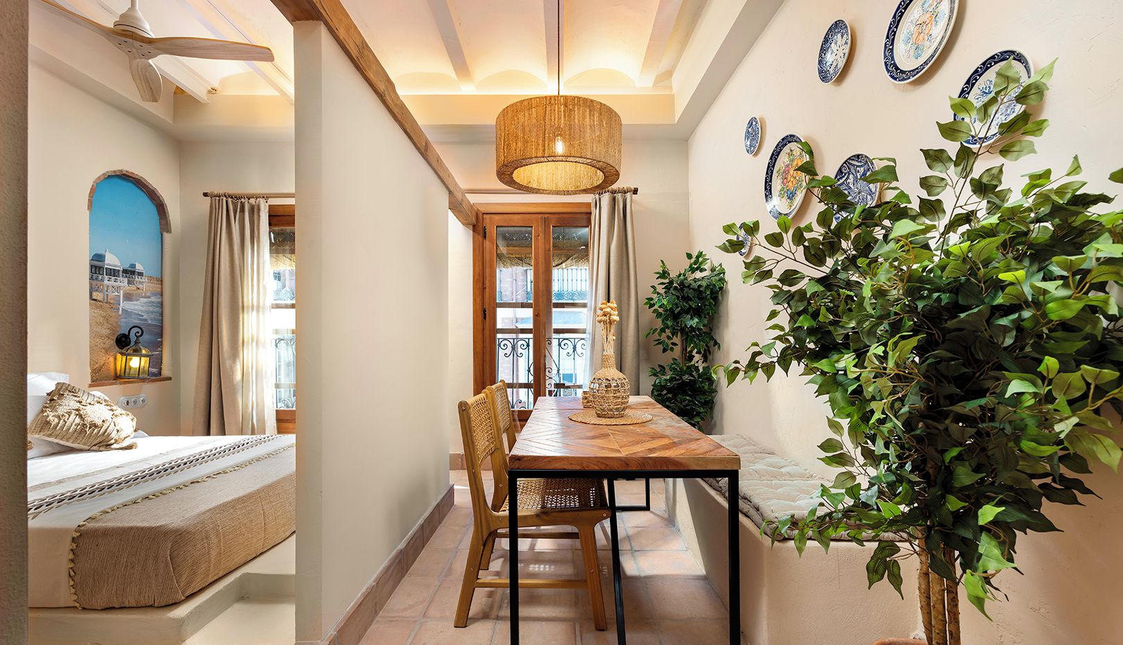 A cozy room with a bed, a dining area with a wooden table and chairs, a potted plant, and decorative plates on the wall.
