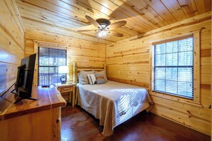 A wooden cabin room with a bed, a desk, a television, and a ceiling fan.