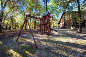 A wooden swing set and climbing structure in a wooded area with a cabin in the background.