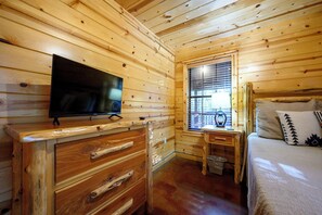 A wooden cabin bedroom with a television, a small table with a lamp, a window with blinds, and a bed with pillows.