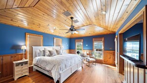 A bedroom with a wooden ceiling, a large bed, a nightstand, a chair, and a window.