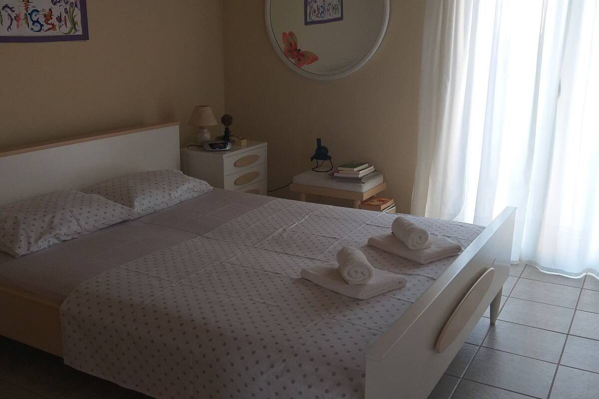 A neatly made bed with a white bedspread and two rolled towels, a bedside table with a lamp, a small shelf with books, and a wall decoration.