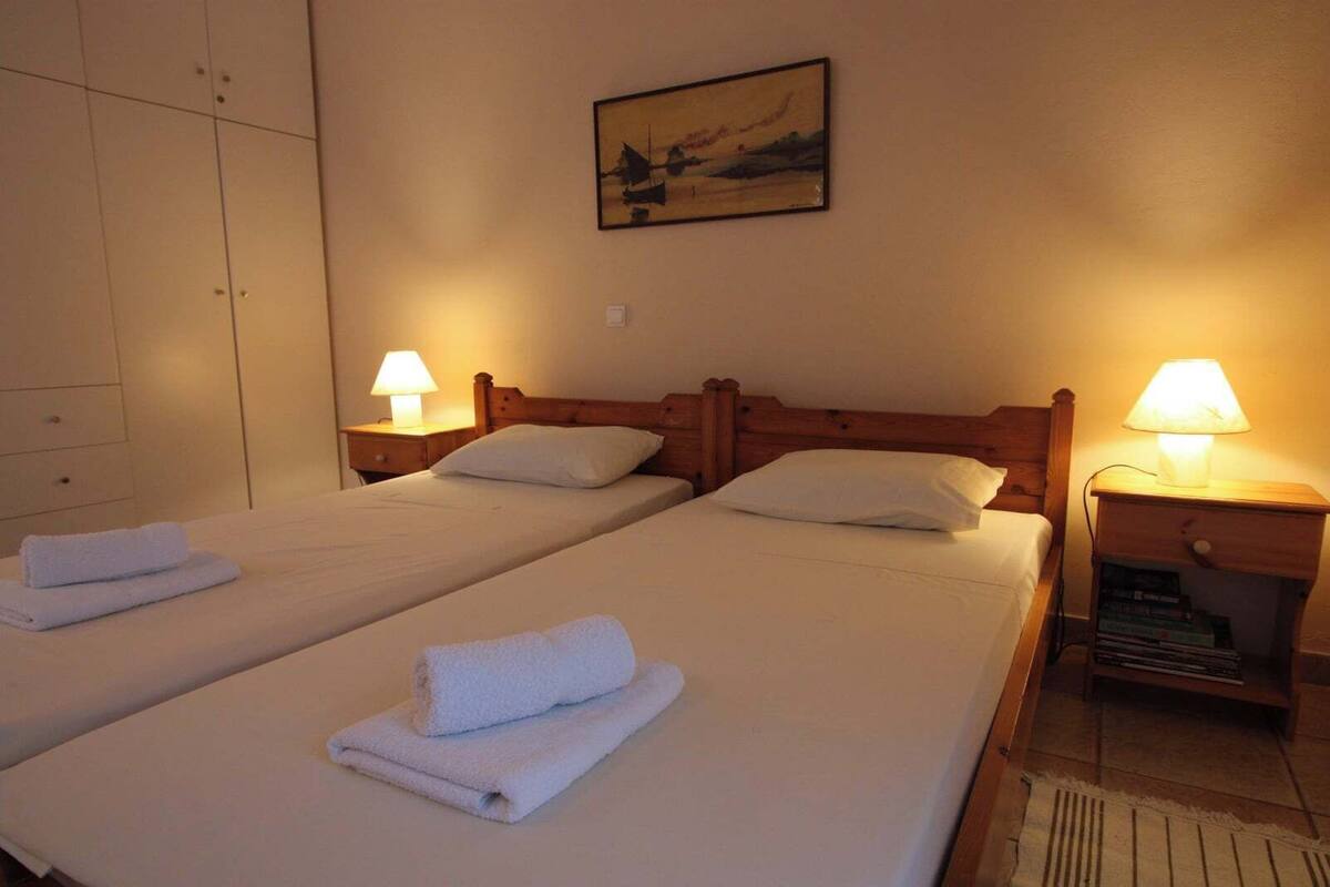 A hotel room with two single beds, each with a wooden headboard, white linens, and a folded towel on the bed. There are two bedside tables with lamps, and a painting hangs on the wall above the beds.