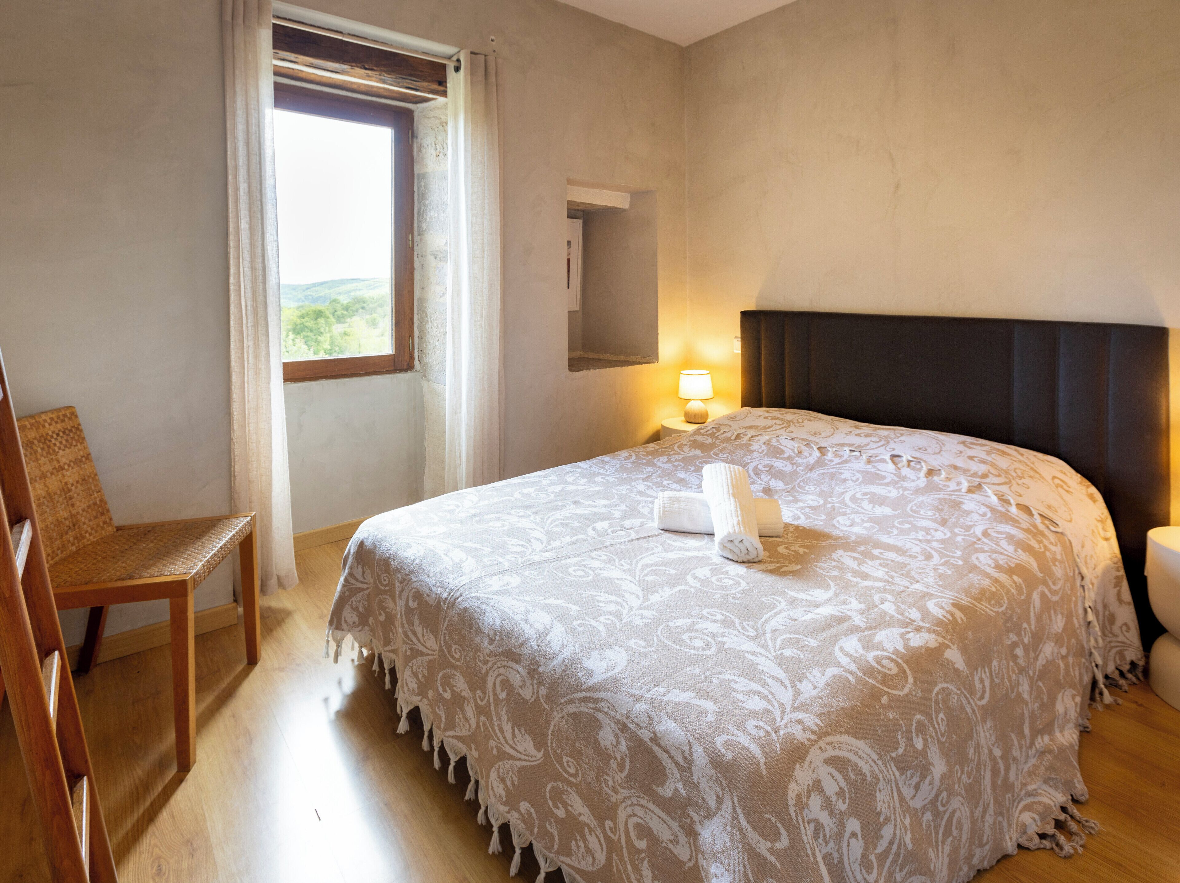 A bedroom with a large bed, a chair, a window with curtains, a bedside lamp, and a view of greenery outside.