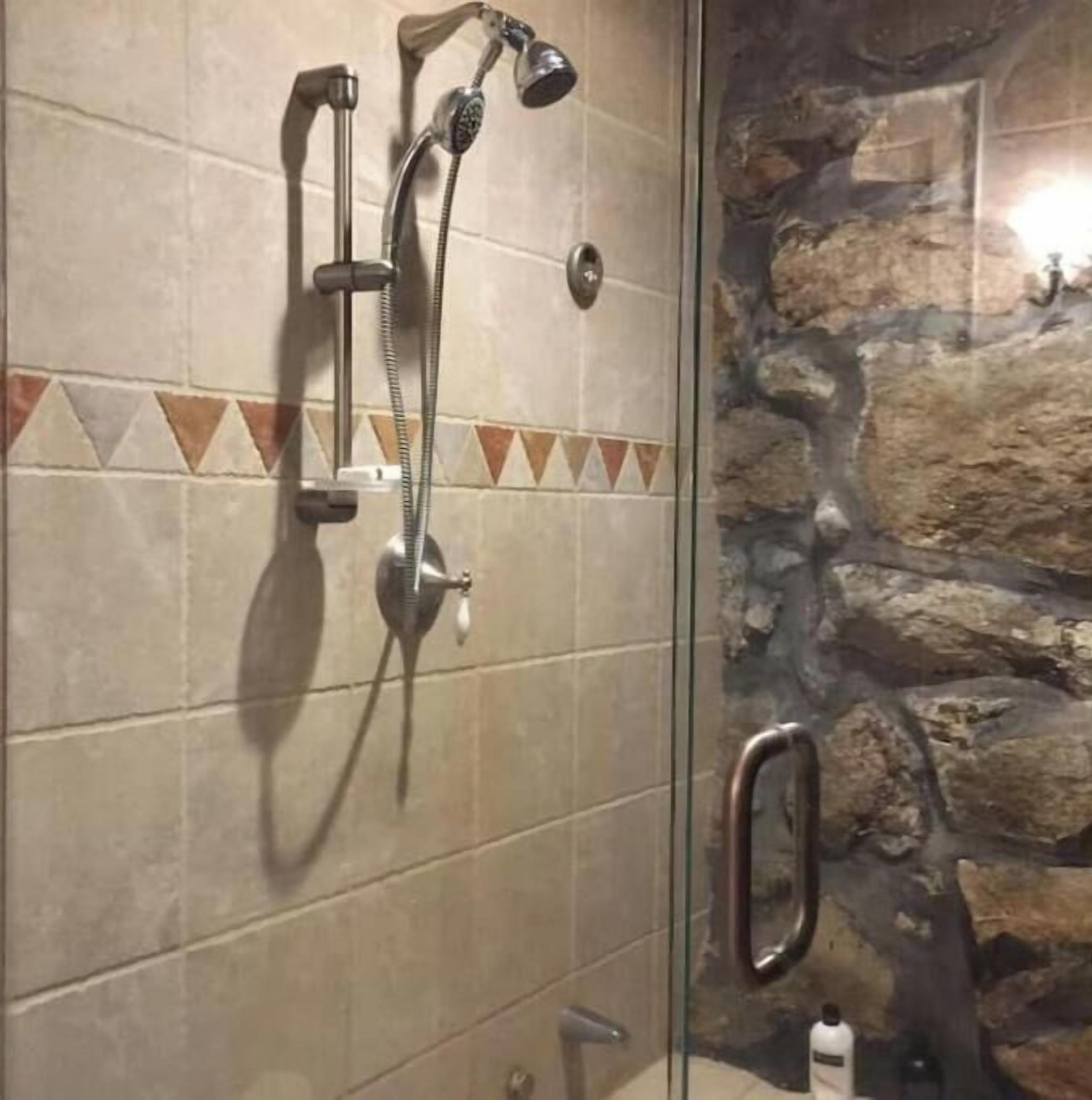 Une douche équipée d’un pommeau de douche à main et d’un robinet mural, d’une porte en verre et d’un mur en pierre.