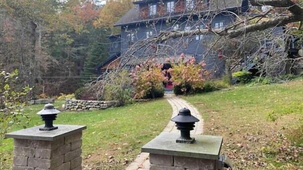 A stone pathway leading to a rustic building with a steep roof, surrounded by autumn foliage and a stone wall.