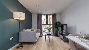 Een moderne woonkamer met een grijze bank, een vloerlamp, een kleine tafel, een televisie, en een eethoek met een witte tafel en stoelen.