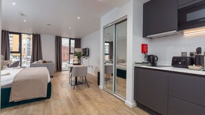 A modern hotel room with a bed, a desk, a chair, a TV, a kitchen area with a microwave, and a sliding glass door to a balcony. - Stylish City Studio (Manchester)