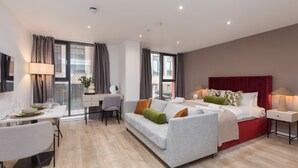 A modern hotel room with a grey sofa, a red headboard, a dining table with chairs, and a large window with curtains. - Spacious City Studio (Manchester)