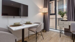 A modern dining area with a flat-screen TV, a table set with plates and glasses, a lamp, and a view of a brick building through large windows. - Spacious City Studio (Manchester)