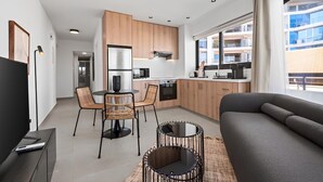 A modern kitchen with wooden cabinets, a dining area with wicker chairs, a sofa, and a large window offering a view of the city. - Finikoudes Marina View 201 - Charming Finikoudes Getaway (Larnaca)