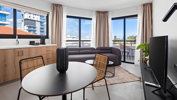 A modern living room with a sofa, a dining table, and a TV. There are large windows with curtains, a kitchen area with cabinets, and a view of buildings outside. - Finikoudes Marina View 201 - Charming Finikoudes Getaway (Larnaca)