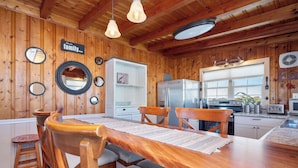 A wooden interior with a dining table, chairs, a refrigerator, and a kitchen area. - Look and Sea: Dog-friendly Ocean View Cottage with Pool Table (Oak Island)