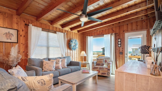 A cozy cabin interior with a wooden ceiling, a ceiling fan, a sofa set, a coffee table, and a view through a door. - Look and Sea: Dog-friendly Ocean View Cottage with Pool Table (Oak Island)