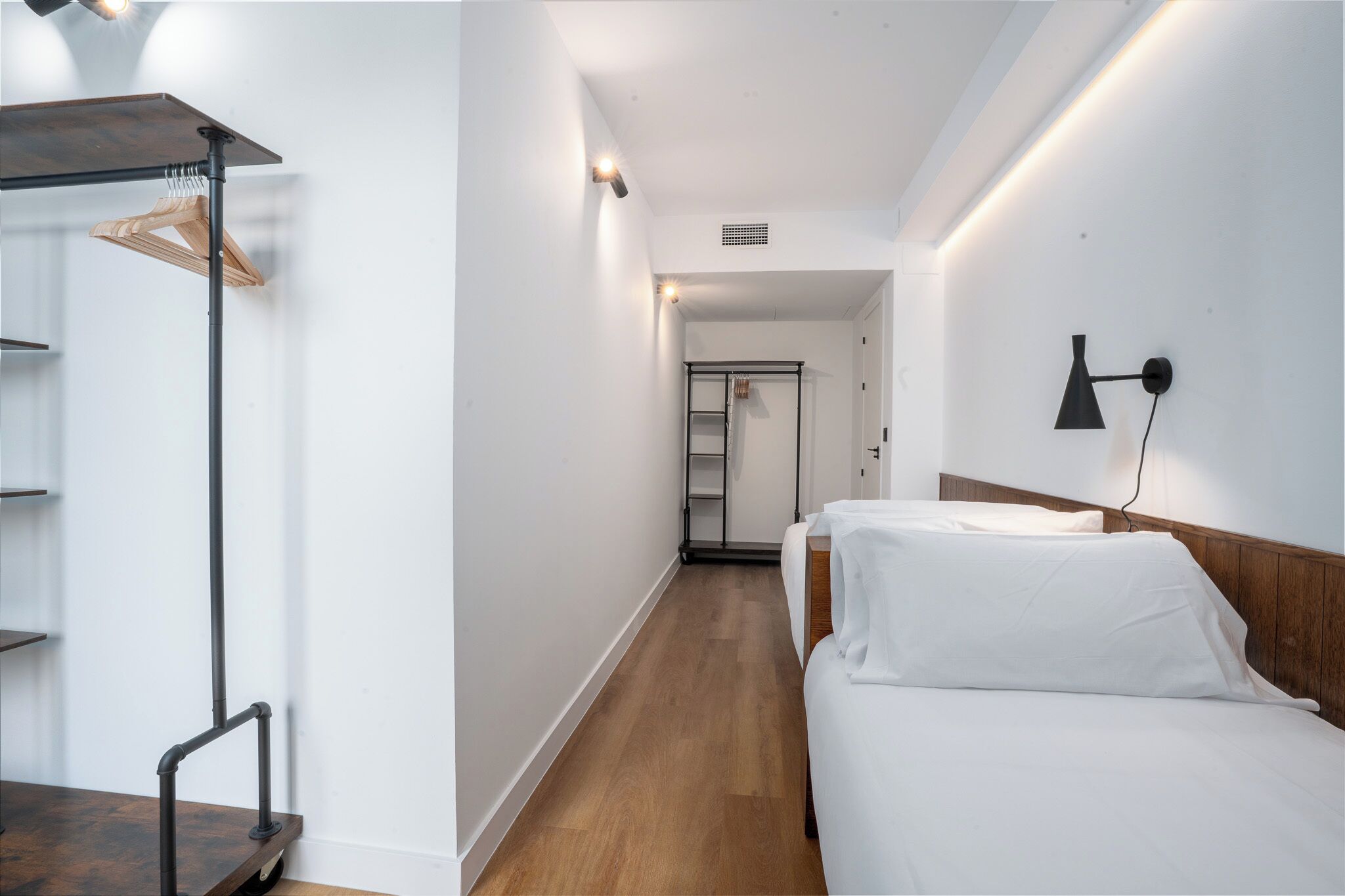 A hotel room with a wooden floor, white walls, a bed with white bedding, a wooden wardrobe, a black wall-mounted lamp, and a hallway leading to a door.
