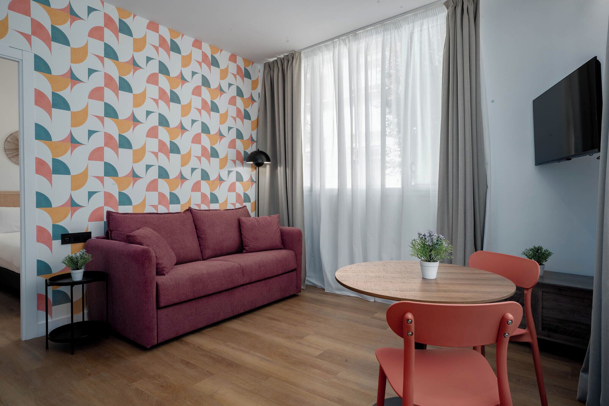 Quarto moderno com parede estampada, sofá, mesa com cadeiras, televisão e janela com cortinas.