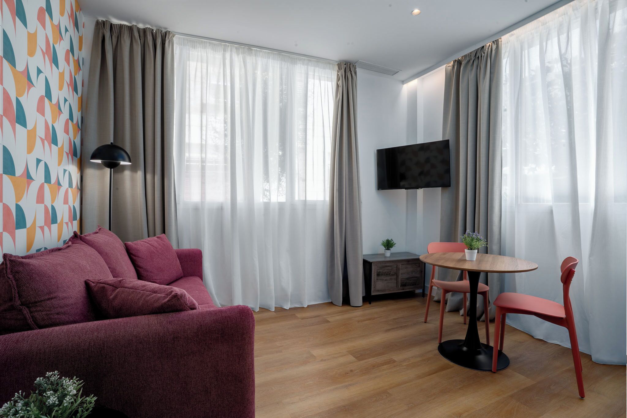 A modern living room with a sofa, a round table with chairs, a TV mounted on the wall, and large windows with curtains.