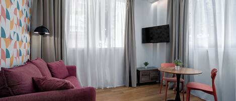 A modern living room with a sofa, a round table with chairs, a TV mounted on the wall, and large windows with curtains.