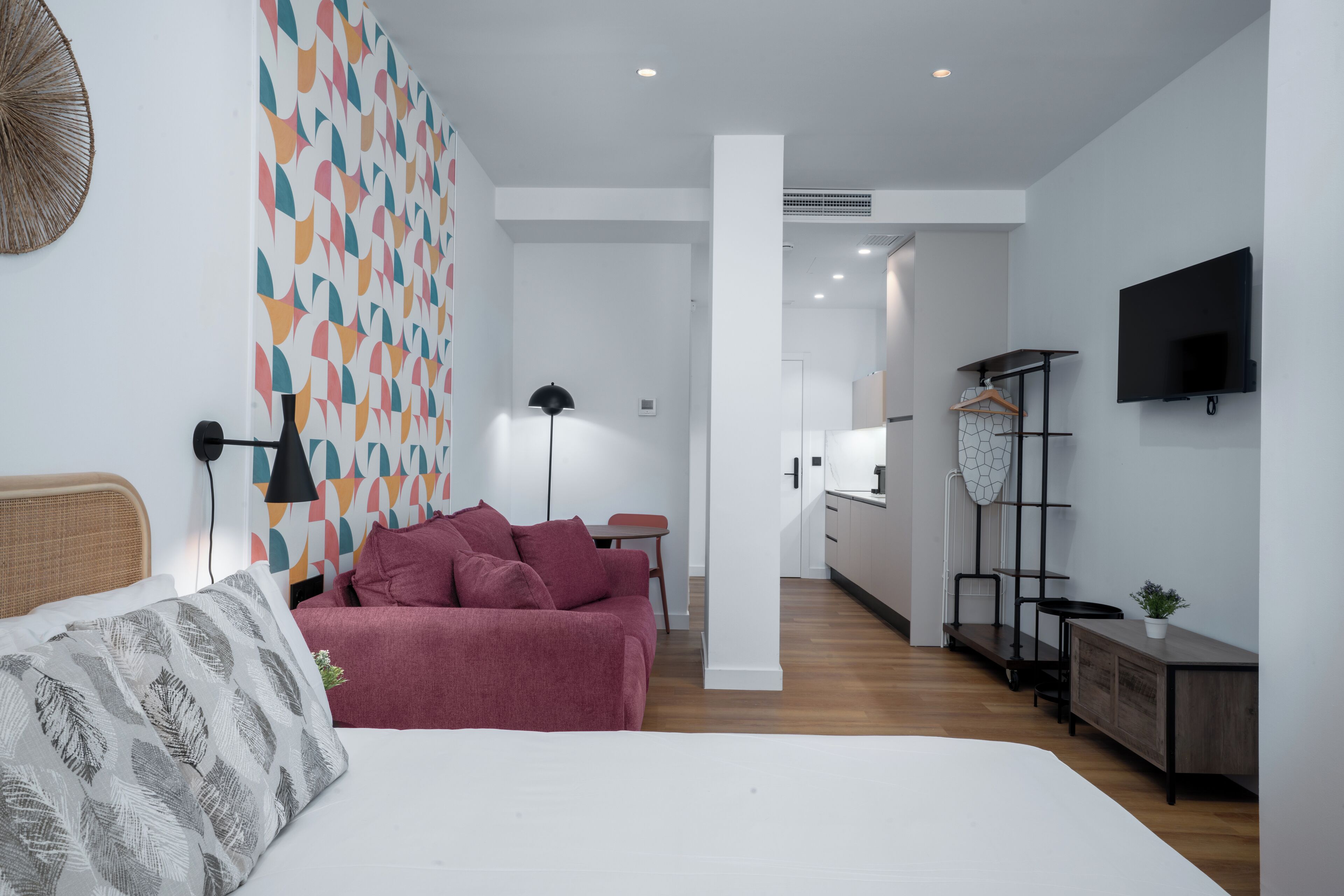 A modern bedroom with a bed, a red sofa, a patterned wall, a TV, and a kitchen area in the background.