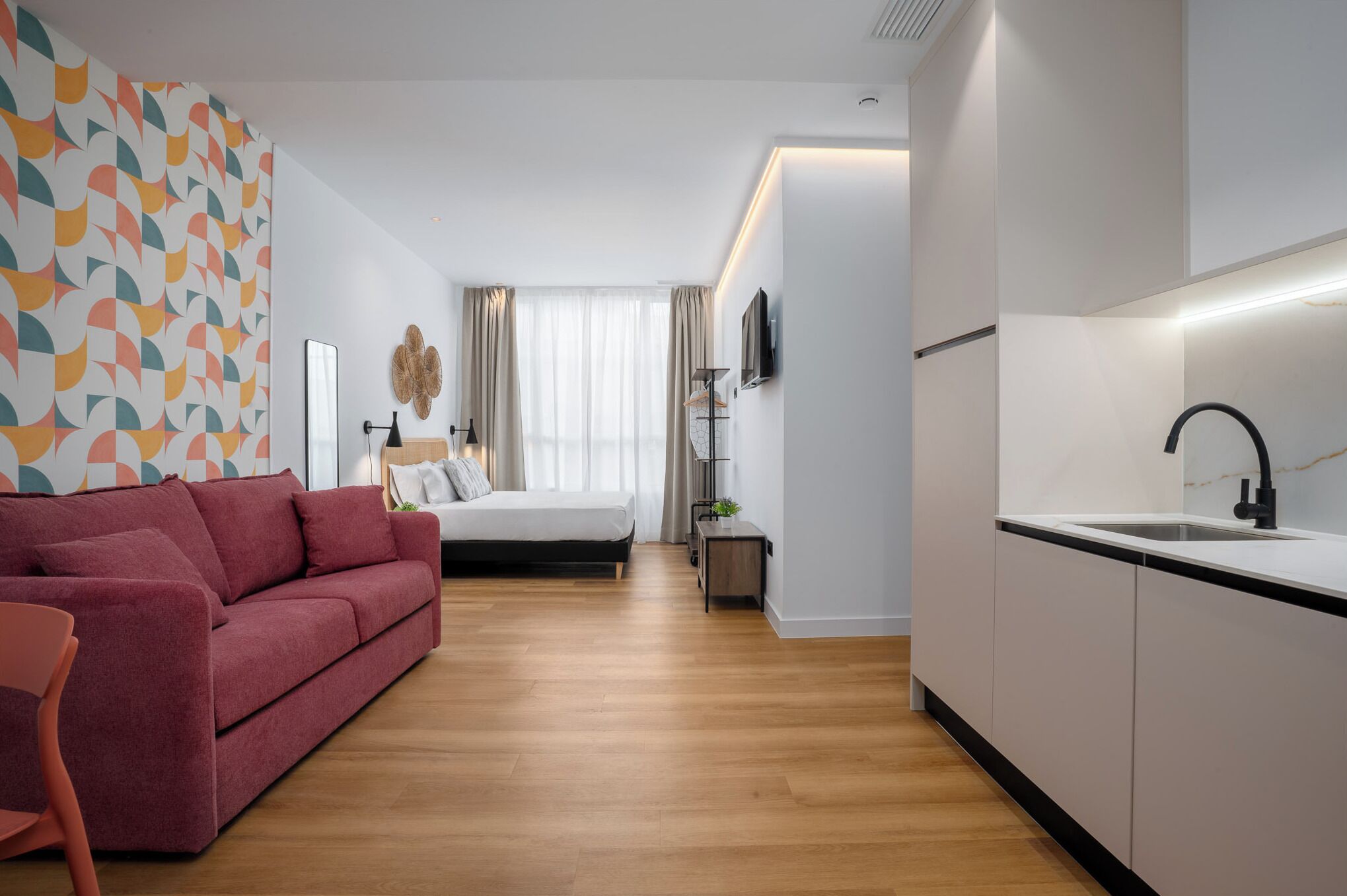 Ein modernes Hotelzimmer mit einem roten Sofa, einem Bett, einem kleinen Tisch mit einer Lampe, einer Küchenzeile mit Spüle und einem großen Fenster mit Vorhängen.