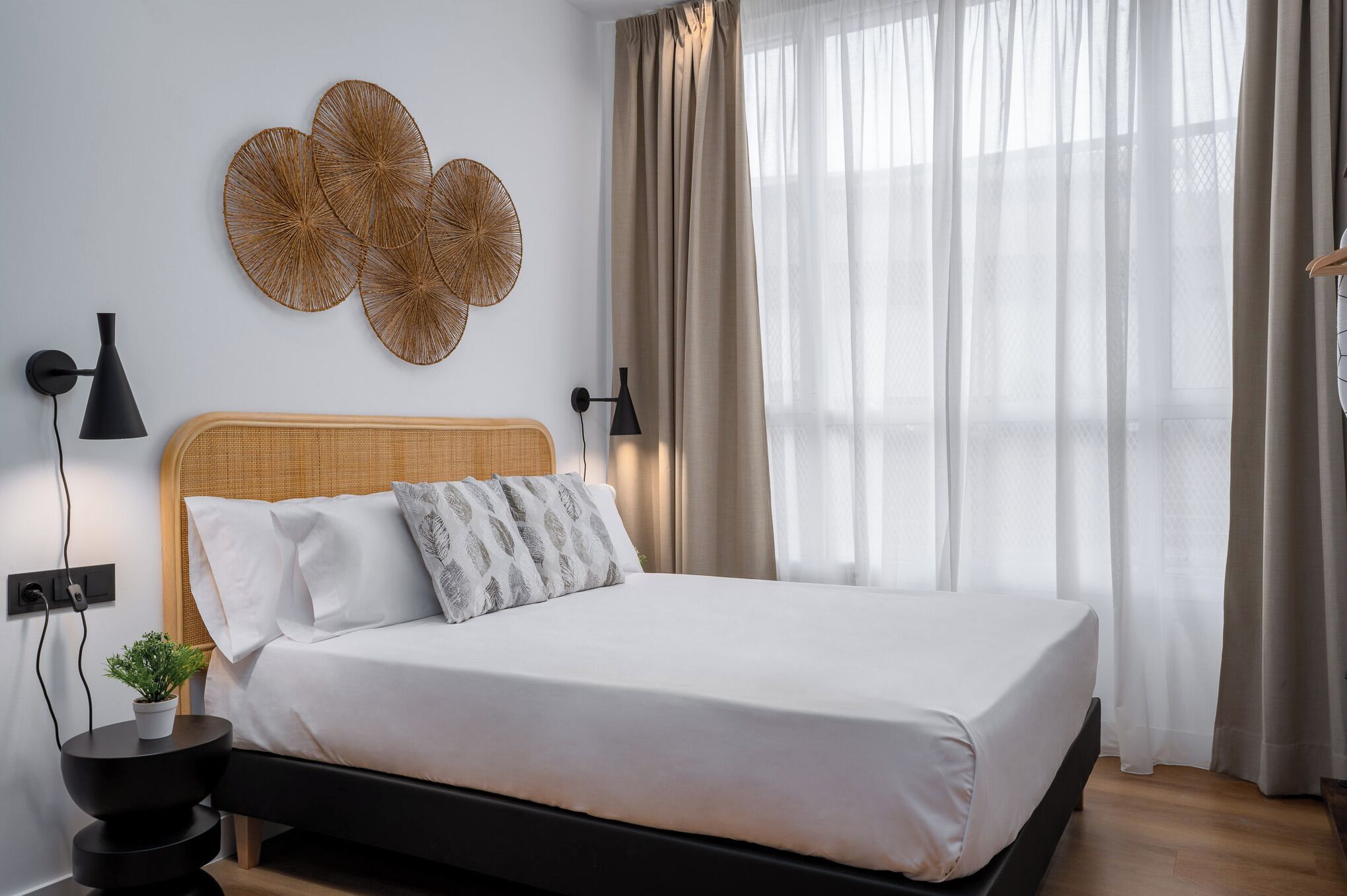 Ein ordentlich bezogenes Bett mit weißer Bettdecke und zwei Kissen, ein gewebtes Wandbild darüber sowie ein Nachttisch mit Lampe und einer kleinen Pflanze.