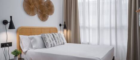 Ein ordentlich bezogenes Bett mit weißer Bettdecke und zwei Kissen, ein gewebtes Wandbild darüber sowie ein Nachttisch mit Lampe und einer kleinen Pflanze.