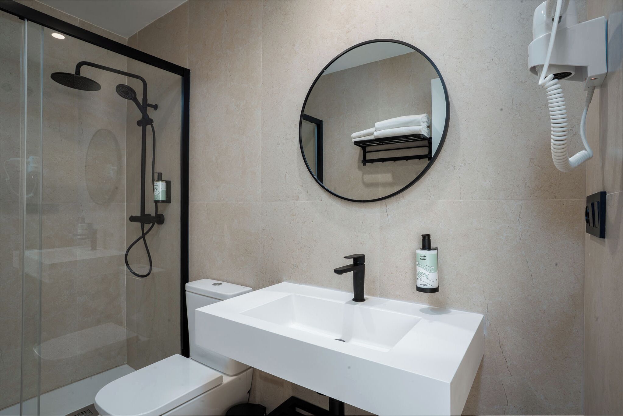 Une salle de bain moderne avec une douche à parois de verre, un lavabo blanc doté d’un robinet noir et un miroir rond placé au-dessus.