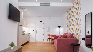 Une chambre d’hôtel moderne comprenant un lit, un canapé rouge, un coin repas avec une table et des chaises, un téléviseur fixé au mur et un mur décoré d’un motif.