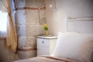 A bedroom with a stone wall, a bedside table with a vase of flowers, a bed with white bedding, and a nightstand with a lamp. - The Palace (Rovinj)