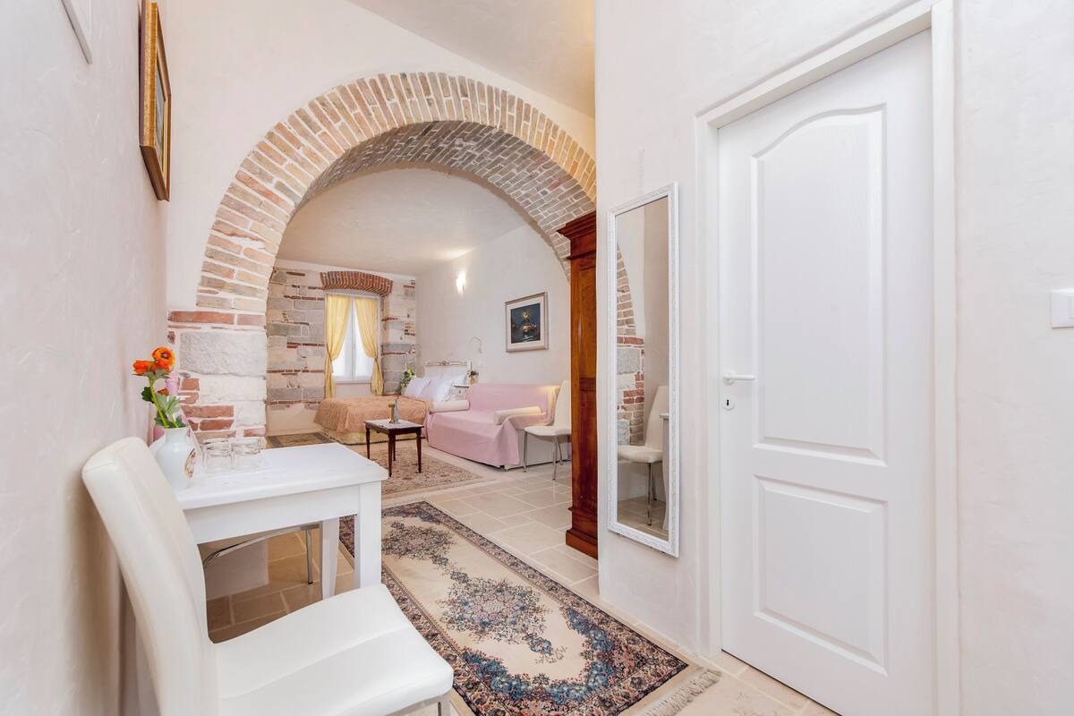 A hallway with a white door, a white table with a vase of flowers, and a brick archway leading to a living room with a pink sofa and a fireplace.