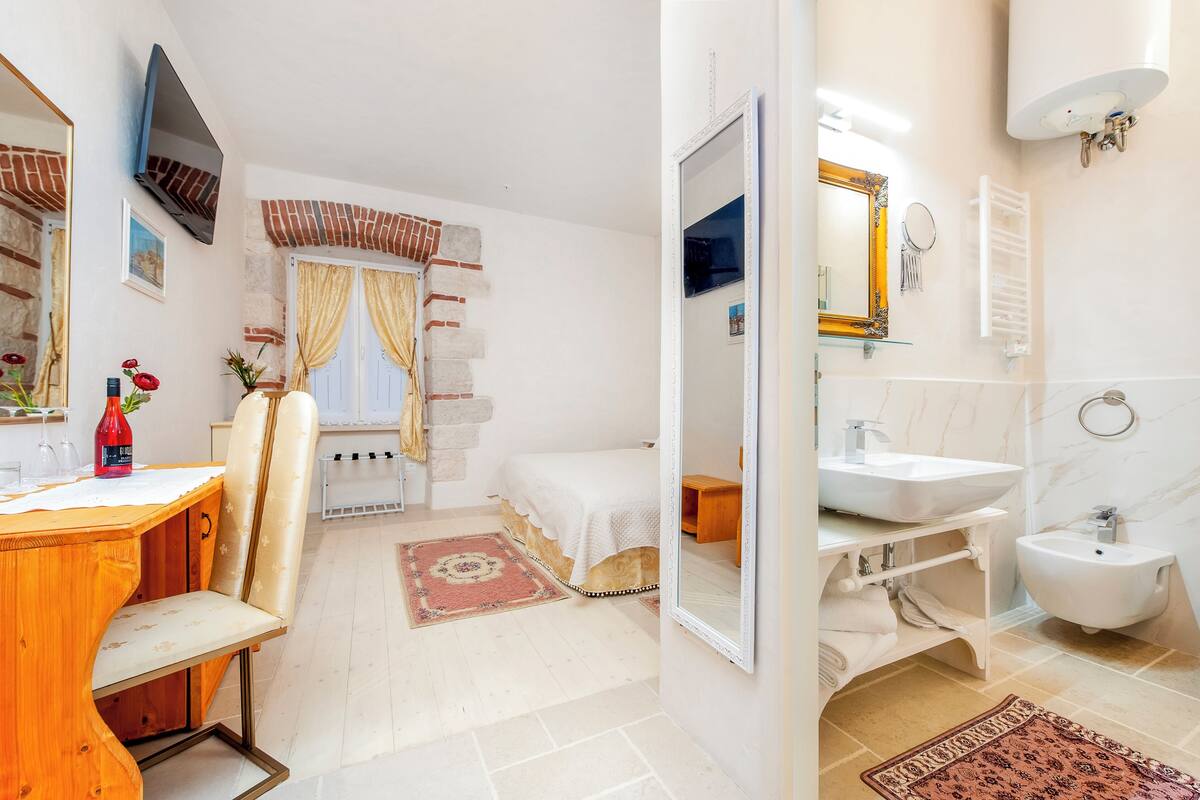 A bedroom with a bed, a wooden desk with a chair, a TV, and a bathroom with a sink, bidet, and mirror.