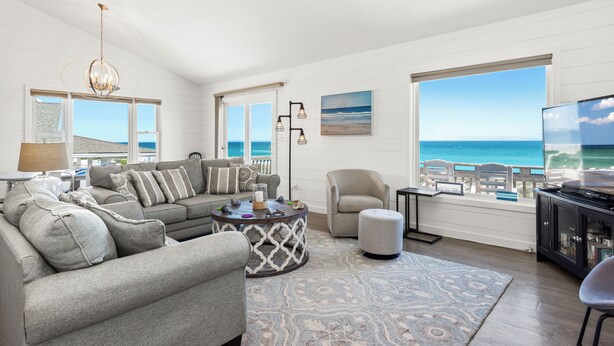 A spacious living room with a large window offering a view of the ocean, a comfortable seating area with a sofa and armchairs, a coffee table, and a television on a stand. - 4 BR Oceanfront Carolina Clipper East has Updated Kitchen with OF Views (Emerald Isle)