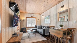 A cozy cabin interior with a wooden ceiling, a fireplace, a sofa, a dining table, and a TV mounted on the wall.