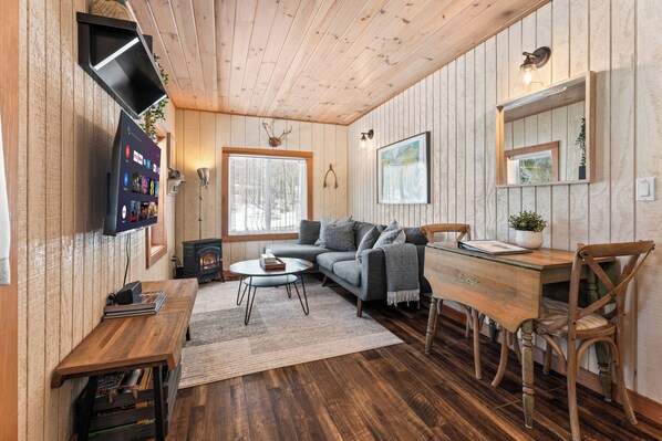 A cozy cabin interior with a wooden ceiling, a fireplace, a sofa, a dining table, and a TV mounted on the wall.