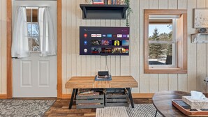 A room with a wooden door, a mounted TV displaying various streaming services, a small table with a laptop, and a window with a view of trees.