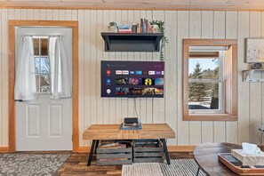 Een kamer met een houten deur, een wandgemonteerde televisie waarop verschillende streamingdiensten worden getoond, een klein tafeltje met een laptop en een raam met uitzicht op bomen.