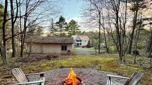 A gravel fire pit with two Adirondack chairs facing it, surrounded by a forested area with houses in the background.