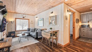 A cozy cabin interior with a fireplace, wooden floors, a sofa, a dining table, and a kitchen area.