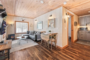 Een gezellig chaletinterieur met een open haard, houten vloeren, een bank, een eettafel en een keukenhoek.
